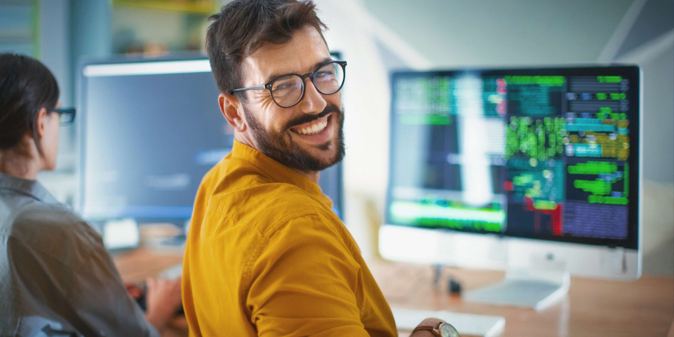Man in yellow cardigan looks back and smiles as he works at computer